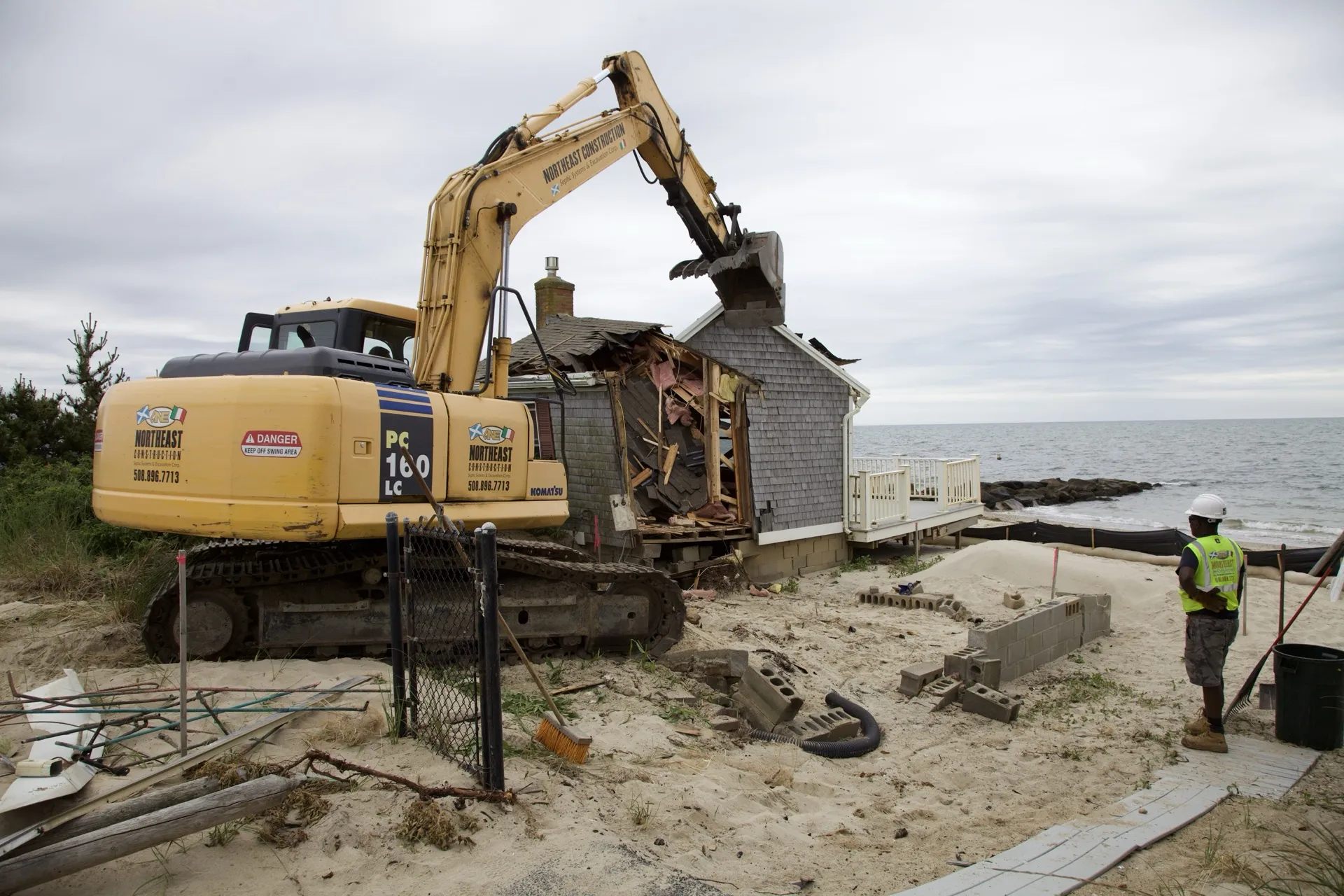 Excavator taking down an aging beachfront cottage in Dennis Port, ocean behind, during Custom Crafted Homes site prep.