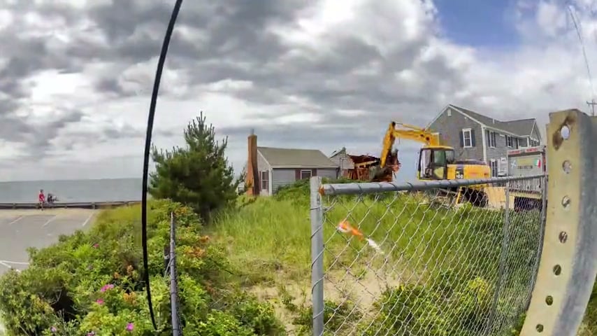 Beachfront home teardown in progress in Dennis Port, MA, with an excavator demolishing the house and the ocean behind