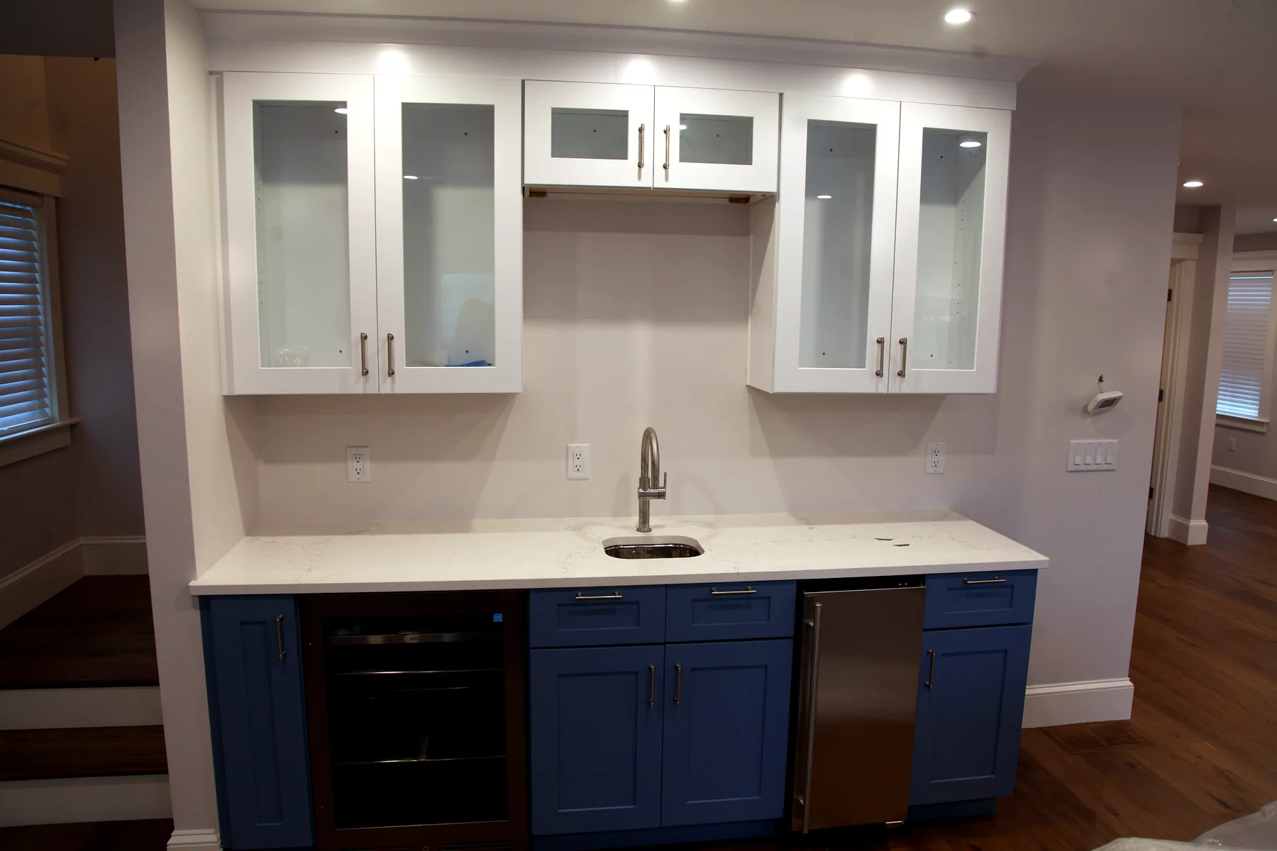 Built-in wet bar with blue lower cabinets, white upper cabinets, beverage cooler, sink, and tile backsplash