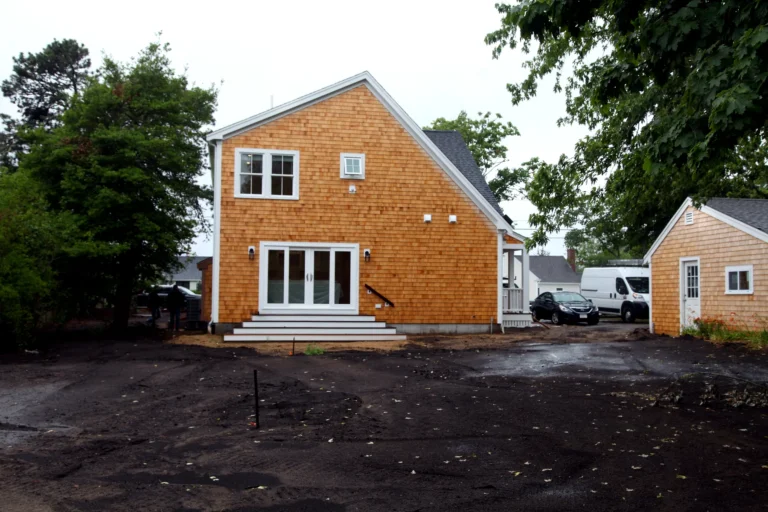 Rear exterior of cedar-shingle Cape Cod home with French doors, detached outbuilding, and wet driveway