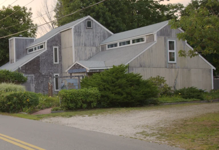 A weathered cedar-shingle and vertical-board building in Wellfleet Center at sunrise, with shed dormers, clerestory windows and steep gable ends.