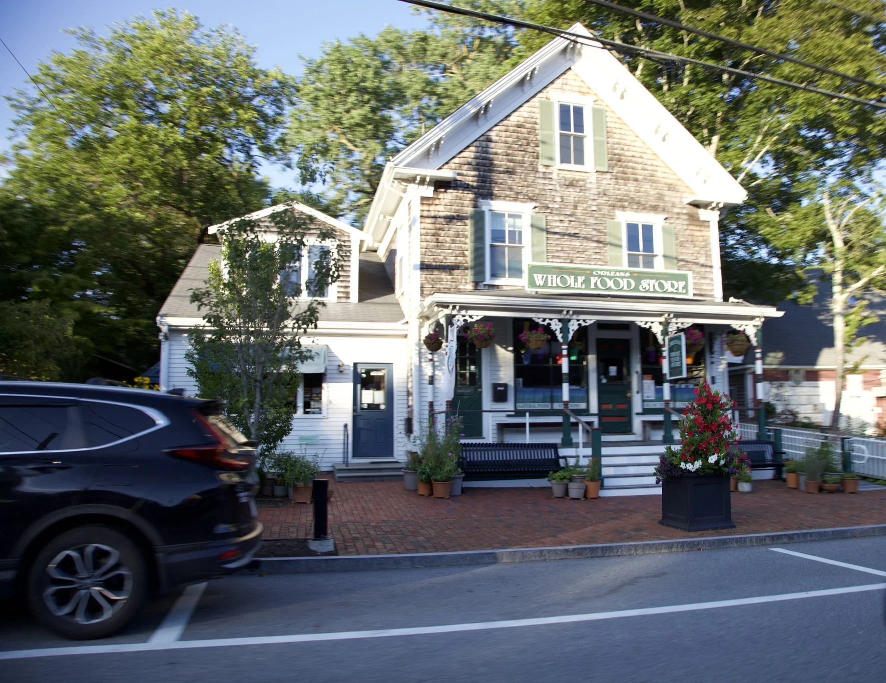 The Orleans Whole Food Store, a cedar-shingled storefront with a covered porch and hanging flower baskets on Main Street in Orleans